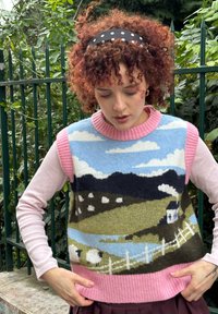Personne aux cheveux roux bouclés et bandeau à pois portant un pull rose et un gilet sans manches en tricot à motif de paysage, debout en plein air.