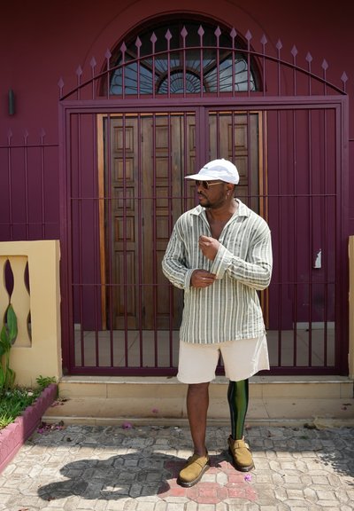 Camisa de rayas de manga larga en verde y crema, pantalones cortos beiges, zapatos marrones y una pierna prostética verde, frente a una pared morada y una puerta de madera.