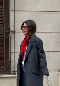 Femme portant un manteau gris foncé, une écharpe rouge, des lunettes de soleil orange, et tenant un sac à bandoulière en chaîne, marchant à l'extérieur près d'un mur beige carrelé.
