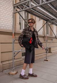 Jachetă maro cu carouri peste un tricou negru, pantaloni scurți gri, șosete albe și pantofi maronii. Ținând o geantă neagră cu un detaliu roșu.
