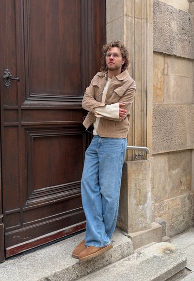 Joven con cabello rizado y gafas apoyado contra una pared de piedra junto a una gran puerta de madera, vestido con chaqueta marrón claro, vaqueros y zapatos marrones.