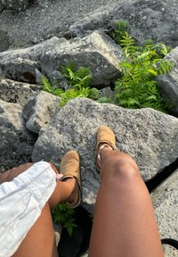 Bruna mockaskor utan snörning på en stenig yta, med grön växtlighet och en vit tygdetalj delvis synlig.