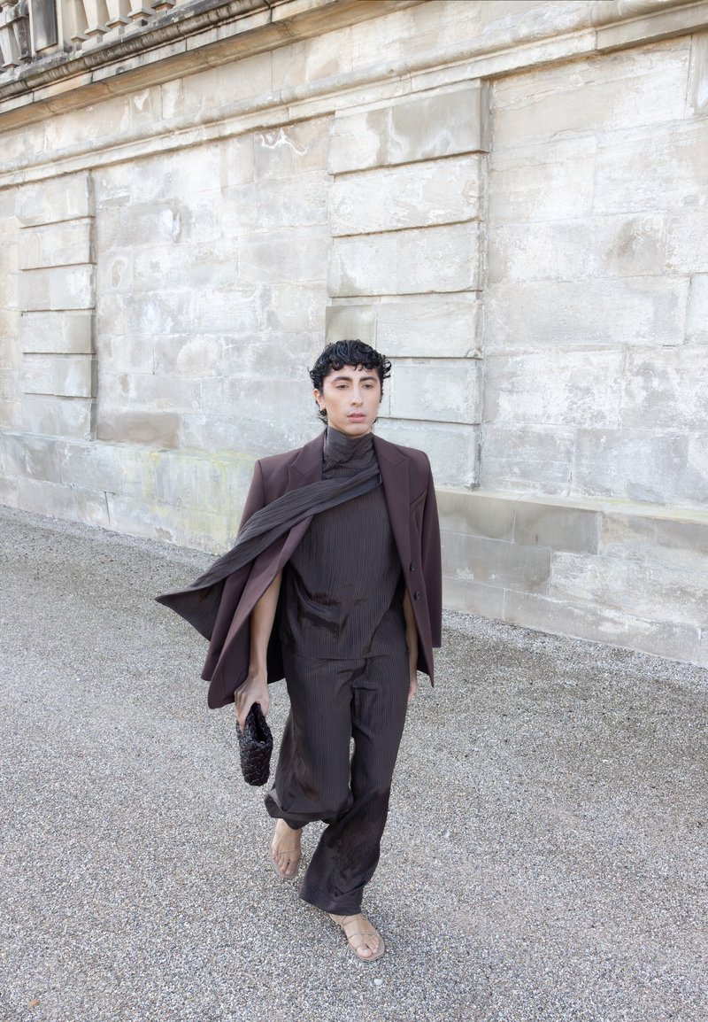 A person walking on gravel beside a stone wall, dressed in a brown suit with a flowing scarf, holding a woven clutch and wearing sandals.