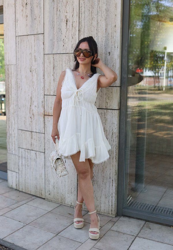 Femme portant une robe blanche sans manches, de grandes lunettes de soleil, tenant un sac à main blanc, debout près d'un mur en pierre et d'une porte en verre.