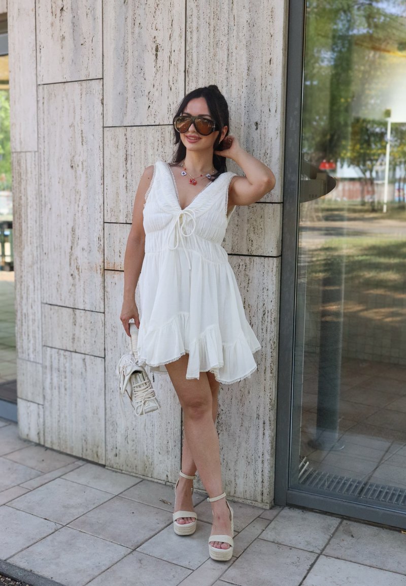 Femme portant une robe blanche sans manches, de grandes lunettes de soleil, tenant un sac à main blanc, debout près d'un mur en pierre et d'une porte en verre.