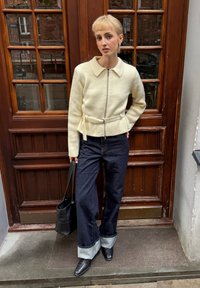 Chaqueta de punto color crema con cierre de cremallera y cinturón, combinada con pantalones anchos de mezclilla oscura con dobladillo vuelto y zapatos negros de punta.