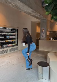 Una persona con una maglia nera a maniche lunghe e jeans ampi blu posa con una gamba sollevata, indossando stivali neri lucidi con tacco alto e fibbia.