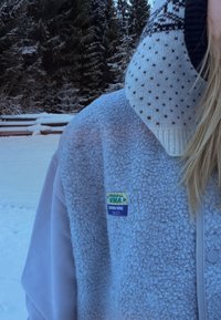 Personne portant une veste polaire bleu clair et un masque facial tricoté à motifs blancs, en plein air dans une forêt enneigée.