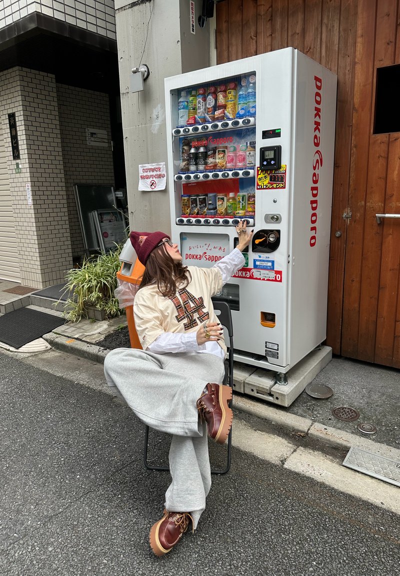 Personne assise sur une chaise portant des vêtements décontractés et un bonnet, tendant la main pour choisir une boisson dans un distributeur automatique blanc Pokka Sapporo.
