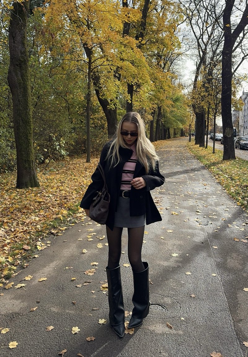 Manteau noir, chemise rayée rose, mini-jupe grise, collants transparents et hautes bottes noires à bout pointu. Sac à main marron, feuilles d'automne en arrière-plan.