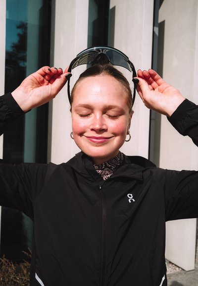 Joven mujer con chaqueta negra sonriendo con los ojos cerrados, levantando gafas de sol oscuras sobre su cabeza al aire libre a la luz del día.