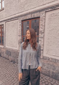 Camisa às riscas azul e branca, tecido leve, design com gola, combinada com calças largas pretas, de pé contra uma parede de pedra.