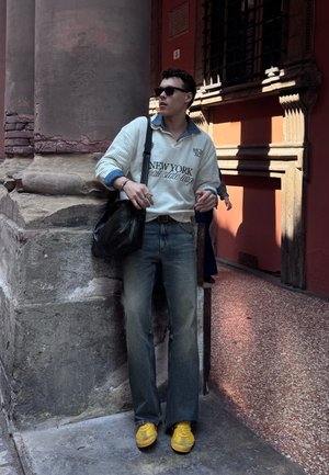 Joven con gafas de sol, suéter blanco, vaqueros y zapatillas amarillas, apoyado contra una columna de piedra en un pasillo histórico iluminado por el sol.
