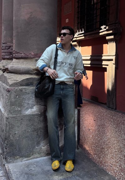 Joven con gafas de sol, suéter blanco, vaqueros y zapatillas amarillas, apoyado contra una columna de piedra en un pasillo histórico iluminado por el sol.