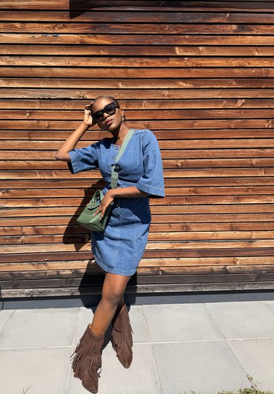 Mujer con gafas de sol, un vestido vaquero, botas marrones con flecos y un bolso bandolera verde se apoya contra una pared de madera con una mano en la cabeza.