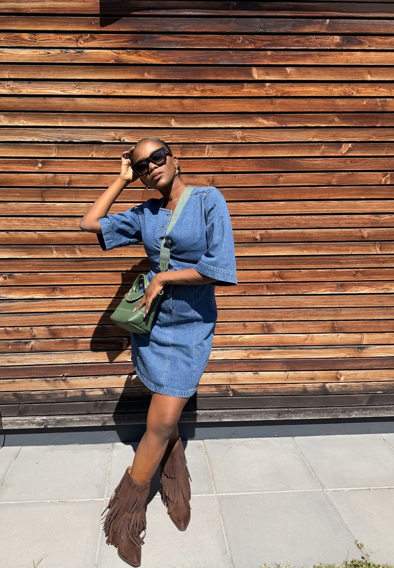 Donna con occhiali da sole, un vestito di jeans, stivali marroni con frange e una borsa a tracolla verde, in piedi contro un muro di legno con una mano sulla testa.