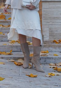 Bež čizme do koljena s izduljenim čarape i uskim siluetom; u kombinaciji s bijelom haljinom koja ima cvjetne čipkaste detalje. Jesenske lišće raspršeno ispod.
