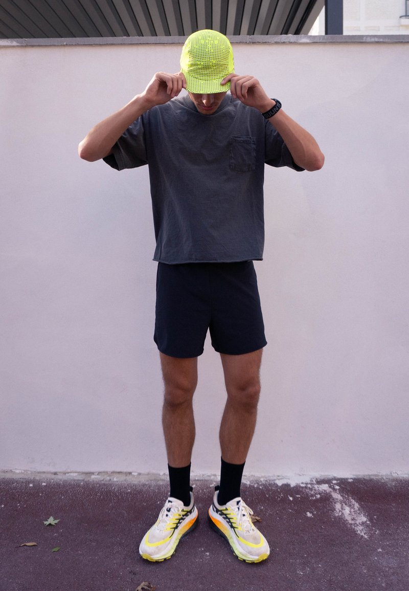 Mann mit neongelber Kappe, dunklem übergroßem T-Shirt, schwarzen Shorts, schwarzen Socken und weißen Laufschuhen steht an einer hellen Wand und richtet seine Kappe.