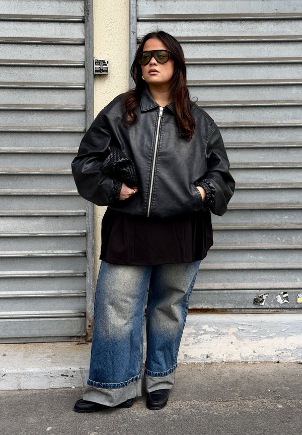 Donna con giacca di pelle nera oversize, jeans a gamba larga risvoltati, scarpe nere, grandi occhiali da sole, che tiene una pochette nera intrecciata davanti a una serranda metallica.