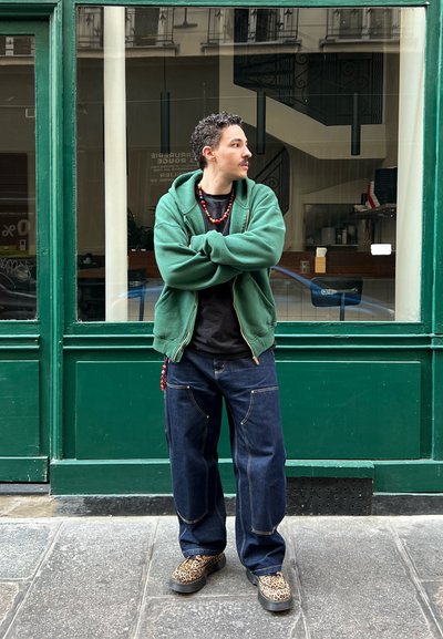 Sudadera con cremallera verde sobre una camiseta negra, combinada con jeans oscuros. El calzado presenta un diseño de estampado de leopardo. El fondo muestra una tienda con fachada verde.