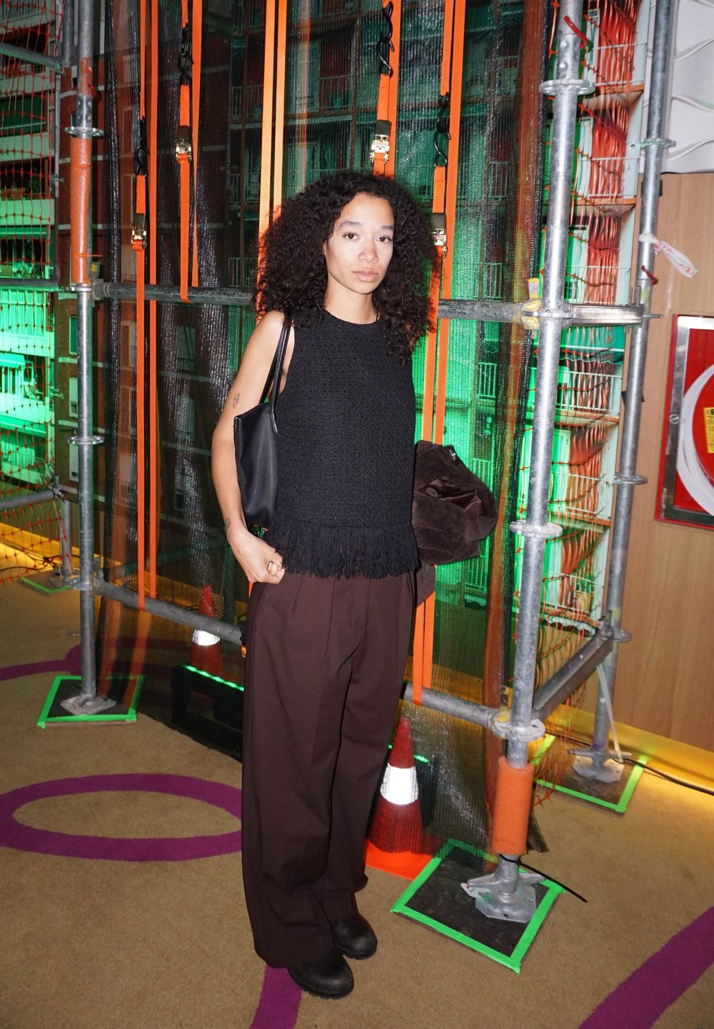 Femme aux cheveux bouclés portant un haut noir sans manches et un pantalon marron, debout près de sangles orange et d'un échafaudage éclairé en vert.