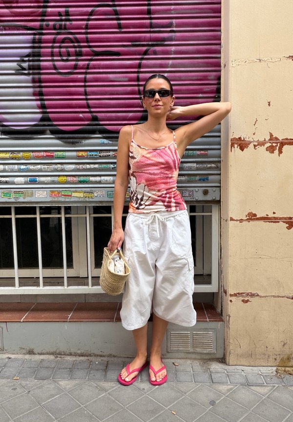 Femme portant des lunettes de soleil et un haut à fleurs se tient devant un volet métallique couvert de graffitis, tenant un sac tissé et portant un pantalon blanc et des sandales roses.