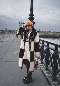 Svart-hvitt rutete fuskepelsfrakk, lagt over en hvit kjole. Tilbehøret inkluderer store solbriller og platåsko med spenner.