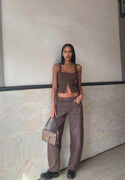 Top de tirantes corto marrón con cierre de botones en la parte delantera, combinado con pantalones anchos de pierna recta marrones. La modelo sostiene un pequeño bolso rectangular.