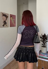 Sieviete ar sarkaniem matiem, ģērbusies melnā svārku komplektā ar uzdūrēm un violetā kreklā ar "93" uz aizmugures, nēsā melnu rokassomu ar uzdūrēm iekštelpās tuvumā mākslai un grāmatām.