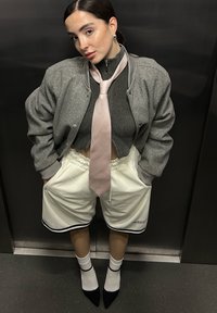 Šedá bombera bunda přes tmavě šedý žebrovaný svetr, bílé síťové kraťasy s černými akcenty, růžová kravata, bílé ponožky, černé špičaté boty.