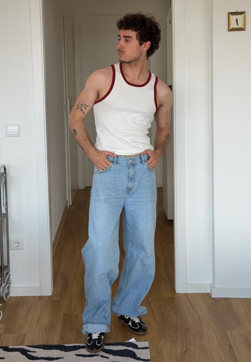 Homme portant un débardeur blanc avec un liseré rouge et un jean bleu ample debout dans une pièce avec un sol en bois et des murs blancs.