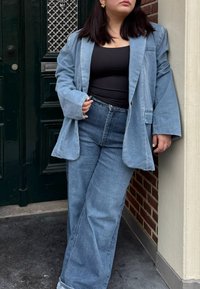 Helleblaue Jeansblazer mit weiten Ärmeln, kombiniert mit einem schwarzen Tanktop und verwaschenen blauen Jeans. Goldene Ohrringe setzen einen auffälligen Akzent.