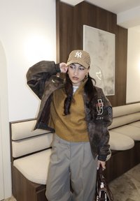 Νεαρή γυναίκα με καφέ μπουφάν, μπεζ παντελόνι και καπέλο μπέιζμπολ με καρό σχέδιο στέκεται μέσα σε χώρο, κρατώντας μια καφέ τσάντα κοντά σε καθίσματα πάγκου.