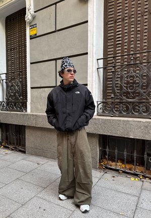 Joven con chaqueta negra, gorro estampado, gafas de sol y pantalones anchos caqui, de pie en la acera de la ciudad junto a ventanas decorativas con barrotes de hierro.