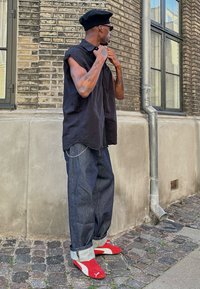 Μαύρο αμάνικο μπλουζάκι, χαλαρά σκούρα τζιν παντελόνια με αναδιπλωμένα μπατζάκια, κόκκινα και άσπρα αθλητικά παπούτσια, φορώντας μαύρο μπερέ και γυαλιά ηλίου.