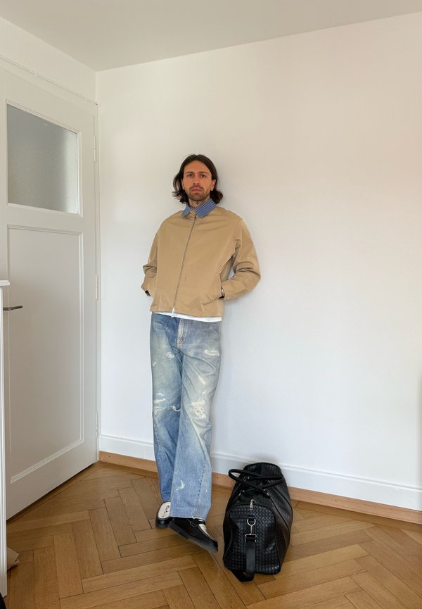 Uomo con capelli alle spalle che indossa una giacca beige, camicia blu con colletto, jeans consumati e scarpe nere, in piedi contro un muro bianco vicino a una borsa da viaggio nera.