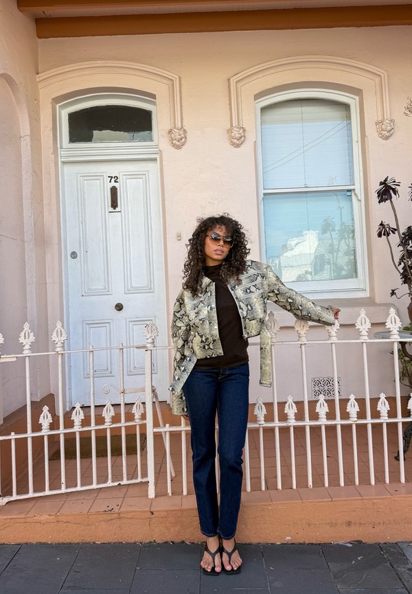 Une femme portant des lunettes de soleil, une veste à motifs et un jean pose près d’une rambarde blanche devant une maison aux tons pastel.