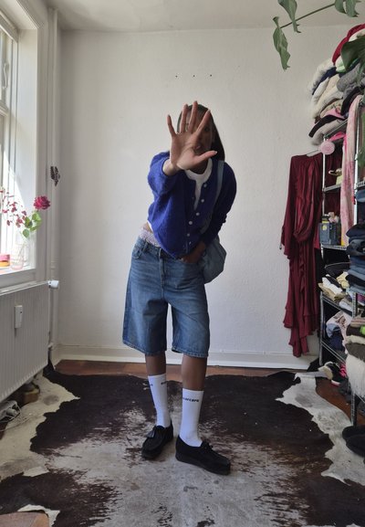 Persona que lleva un cárdigan azul, pantalones cortos vaqueros, calcetines blancos y zapatos negros, está de pie en el interior, sosteniendo una bolsa con una mano y cubriéndose la cara con la otra.
