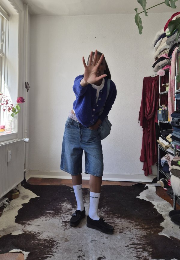 Osoba odjevena u plavi kardigan, traper kratke hlače, bijele čarape i crne cipele stoji u zatvorenom prostoru, držeći torbu jednom rukom i pokrivajući lice drugom.