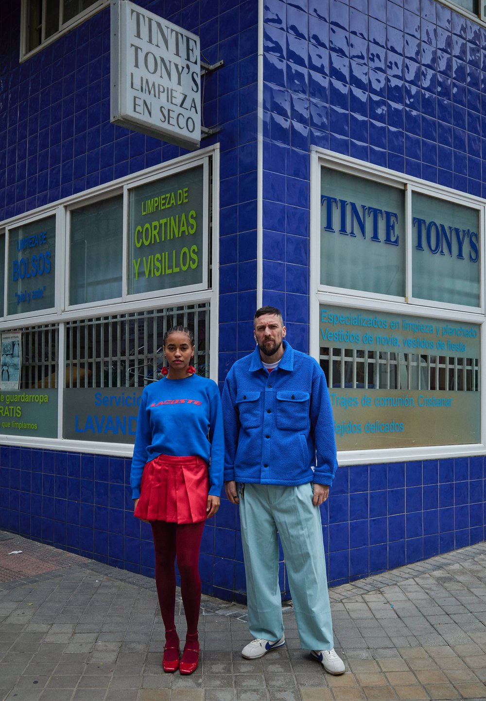 Edifício com azulejos azuis com a placa "Tinte Tony's". Dois modelos estão do lado de fora a usar roupas coloridas: saia vermelha, suéter azul, calças claras e sapatos.