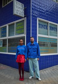 Edifício com azulejos azuis com a placa "Tinte Tony's". Dois modelos estão do lado de fora a usar roupas coloridas: saia vermelha, suéter azul, calças claras e sapatos.