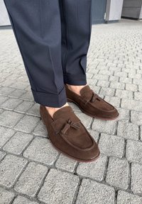 Mocasini maro din piele întoarsă cu ciucuri, purtați cu pantaloni bleumarin înfășurați, pe o pavea gri de piatră cubică, în aer liber.