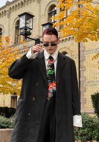 Manteau en laine noire avec de grands boutons, porté sur une chemise blanche. Comprend une cravate colorée à motifs. Lunettes de soleil foncées complètent le look.