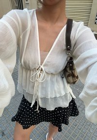 Valkoinen läpikuultava paita, jossa on solmimisosa edessä ja kerroksellinen muotoilu; yhdistetty mustiin pilkkuhameeseen. Korostettu ruskealla olkalaukulla.