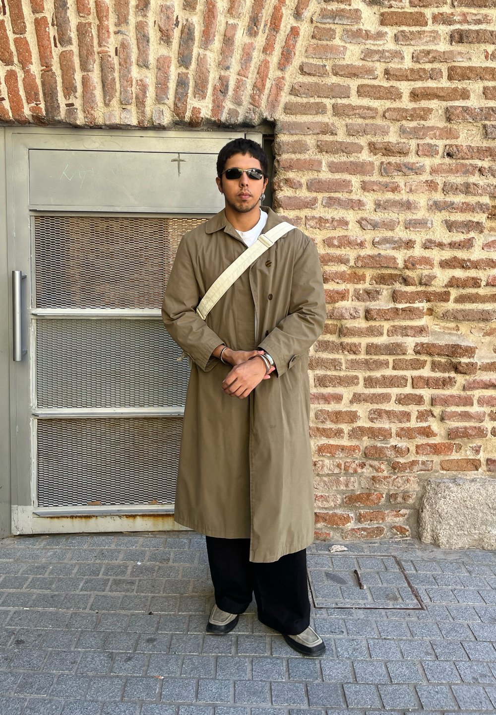 Μακρύ, ελαιοπράσινο παλτό, μαύρο παντελόνι και γκρι παπούτσια. Στηρίζεται σε μια υφαντή τοιχοποιία από τούβλα, φορώντας γυαλιά ηλίου και τσάντα ώμου.