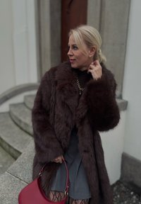 Manteau en fausse fourrure marron avec col châle, porté avec un haut foncé, une jupe grise et un sac à main en cuir rouge. Accessoires inclus : boucles d'oreilles dorées.