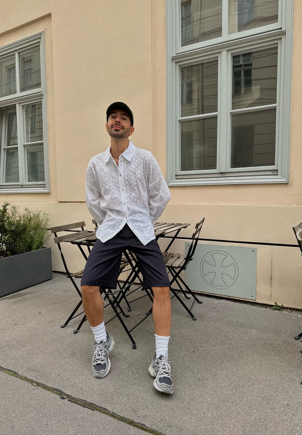 Camisa blanca de botones texturizada, pantalones cortos de rayas marino, zapatillas grises y calcetines blancos; sentado en una mesa de bistró contra una pared clara.