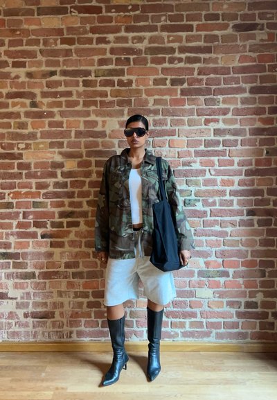 Chaqueta de camuflaje, camiseta sin mangas blanca, pantalones cortos grises, bolso negro de mano y botas negras de tacón alto hasta la rodilla, frente a un fondo de pared de ladrillo.