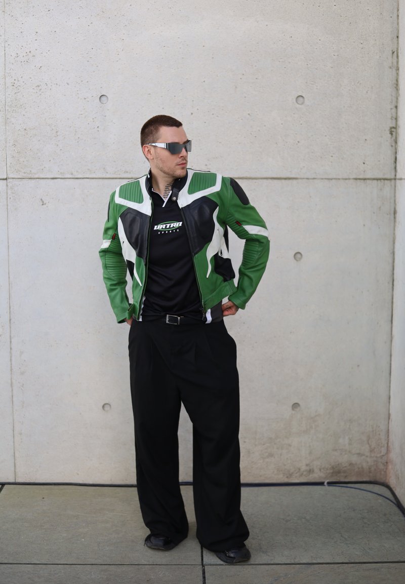 Πράσινη και μαύρη υφασμάτινη δερμάτινη τζάκετ με λευκές λεπτομέρειες, κλείσιμο με φερμουάρ, φορεμένη πάνω από μαύρο πουκάμισο, συνδυασμένη με μαύρο παντελόνι και παπούτσια.