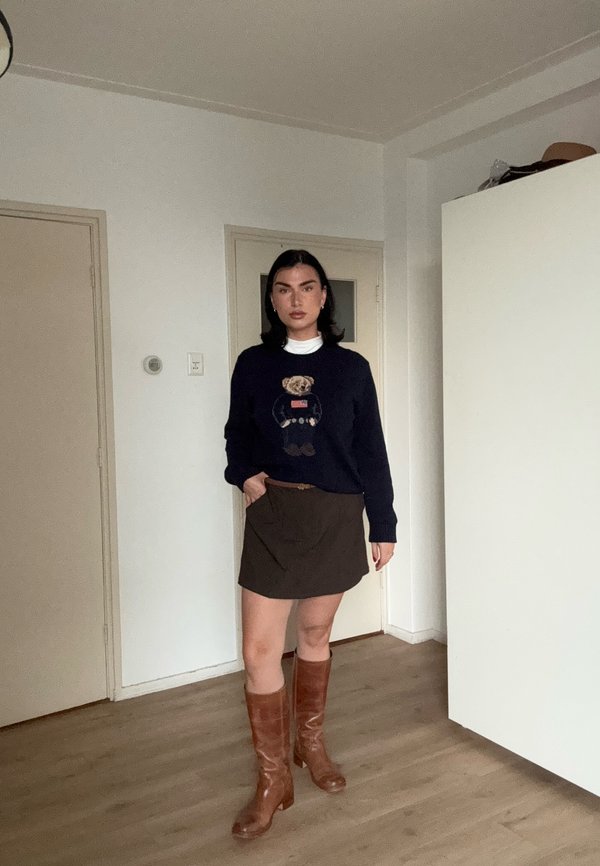 Una mujer posa en interiores con un jersey azul marino, una minifalda marrón y unas botas marrones hasta la rodilla.